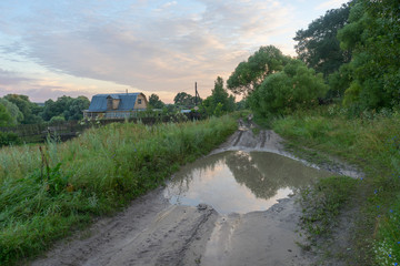 Fototapeta premium Vanishing dirt road with deep rut and puddles in village at sunset