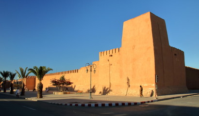 Wysoki, poteżny mur marokańskim mieście, dekoracynie wykończony, z gliny pise, na ulicy kolkoro mieszkańców, lampy uliczne, palmy, intensywnie niebieskie niebo - obrazy, fototapety, plakaty