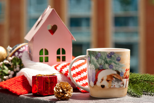 New Year's tea in the morning on the balcony. A mug of tea with a picture of a dog in a santa hat, a toy house and spruce branches create a mood for the new year.