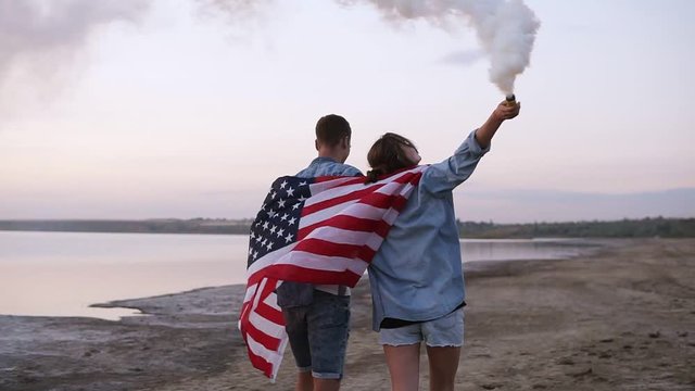 Young people walking outdoors. Young man and woman covered with American flag, going by seashore. Girl in casual walking with a smoke bomb, raised it up