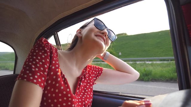 Happy girl in sunglasses leaning out of retro car window and enjoying trip. Young woman looking out window of moving old vintage auto on sunny day. Travel and freedom concept. Slow motion Close up