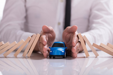 Businessperson Protecting Blue Car From Falling Wooden Dominos