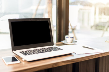 Fototapeta premium Mockup image of laptop with blank black screen,smartphone,coffee cup on wooden table of In the coffee shop.