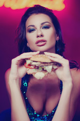 Sexy gorgeous woman in night dress with sequins eating hamburger in night club. Girl standing and posing against red wall with neon letters