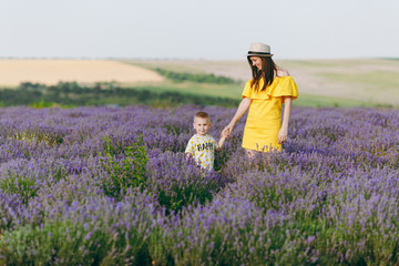 Young woman in yellow dress walk on purple lavender flower meadow field background, rest, have fun, play with little cute child baby boy. Mother, small kid son. Family day, parents, children concept.
