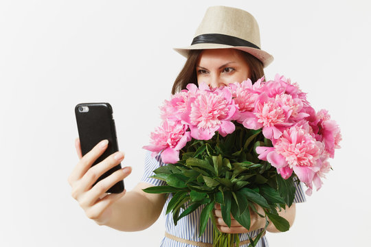 Young woman in blue dress, hat holding bouquet of beautiful pink peonies flowers, doing selfie on mobile phone isolated on white background. St. Valentine's, International Women's Day holiday concept.