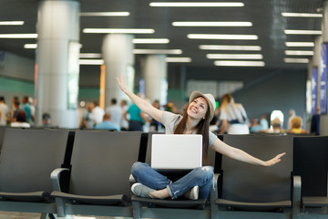 Young dreamy traveler tourist woman with laptop sit with crossed legs spreading hands as in flight, waiting in lobby hall at airport. Passenger traveling abroad on weekend getaway. Air flight concept.