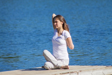 Young caucasian woman doing yoga asanas outdoors on a wooden pier by the blue water of a river or lake, sunny warm summer weather, natural face with no makeup, active and healthy lifestyle concept
