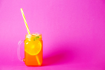 Colorful composition with vintage mason jar glass full of fresh orange juice based non alcoholic summer cocktail with ice, straw, fruit, isolated on pink background. Close up, copy space, top view.