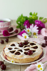 A cherry pie decorated with heart-shaped ornamentation with a cup of tea, a bouquet of flowers and ripe cherry berries.