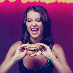 Sexy gorgeous woman in night dress with sequins eating hamburger in night club. Girl standing and posing against red wall with neon letters