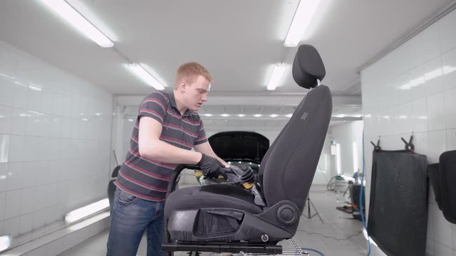 redhead man is hoovering black car seat in a room of auto-service during cleaning, car washing