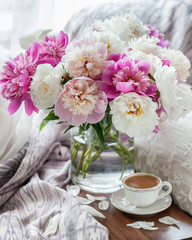 Glorious pastel bouquet of peonies in glass vase on table by window. Cup of coffee. Soft focus.