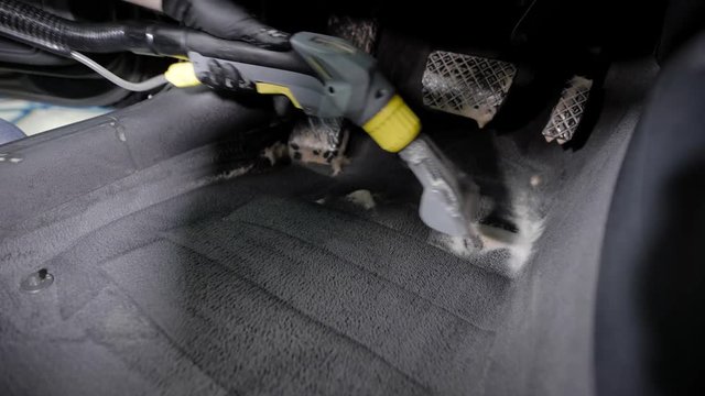 washerman is hoovering automobile carpet, cleaning by foam interior of car in a garage, wearing gloves