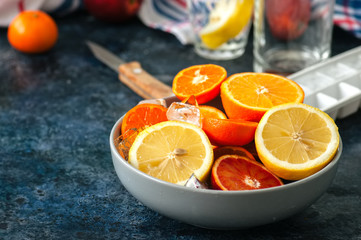 Mix of fresh ripe citrus fruits as blood oranges, mandarines, lemons  with ice cubes in a bowl on a blue stone background.