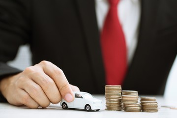 Business man in suit and red necktie holding model of toy car on over a lot of stacked coins - insurance, loan and buying car concept	