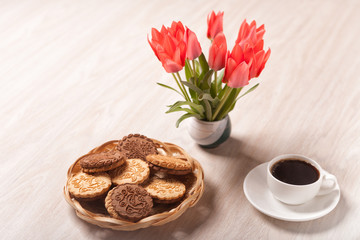 Cookies in a wicker basket, white Cup with coffee on a saucer and a bouquet of tulips on a light textured wooden surface