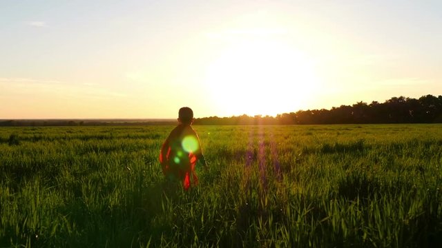 A child in a superhero costume in a red cloak runs across the green lawn against the backdrop of the sunset.