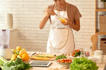 Concentrated Asian food blogger wearing apron using tweezers while putting slices of fresh fruits in container, interior of modern kitchen on background