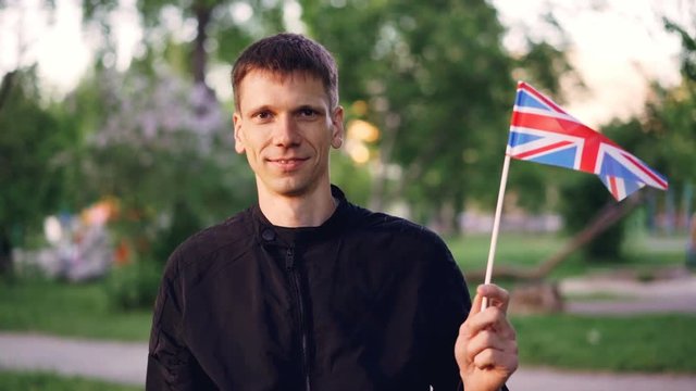 Slow motion portrait of smiling young man proud citizen holding British flag standing in the streen with nice park in background and looking at camera.