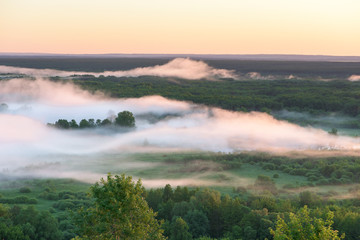 dense fog creeps over the trees near the river at dawn