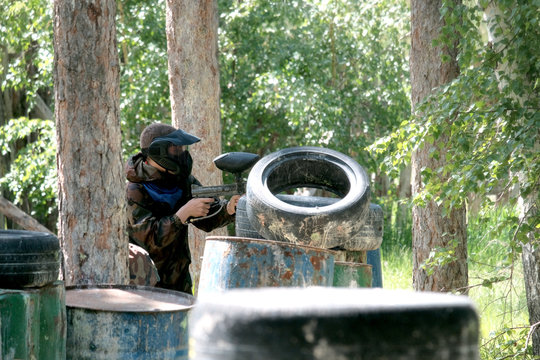 A guy in camouflage clothes from a blue team with weapons in his hands peeks out from behind an iron barrel and tires. Sport game in paintball.