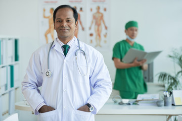 Portrait of Indian doctor standing at his office with male nurse in the background