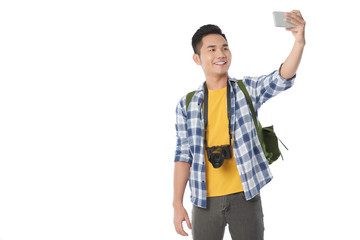 Cheerful Asian tourist with backpack taking selfie on modern smartphone while standing against white background, copy space