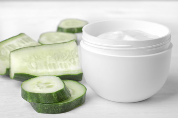 Face cream in a white jar with cucumber extract next to a set of fresh cucumbers on a white wooden background