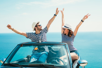 Young attractive couple posing in a convertible car, by the sea - obrazy, fototapety, plakaty