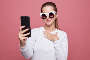 Cheerful fashionable hipster lady in sunglasses has appealing appearance, holds modern smart phone, glad to receive message from best friend, reads invitation on party, rejoices good relationships.