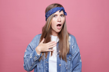 Perplexed indignant female with hippie look, keeps mouth opened, raises eyebrows, opens mouth,points at herself isolated over pink background. Annoyed irritated Caucasian young woman poses in studio.