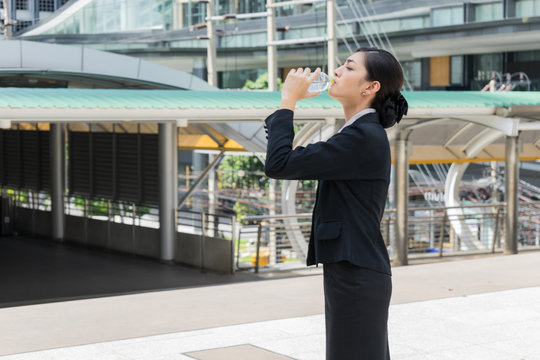Asian businesswoman standing and drinking pure mineral water from plastic bottle with background of building.