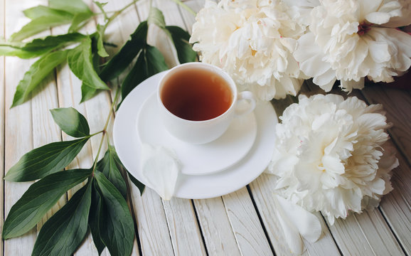 Cup of morning tea and white peonies on light gray wooden background. Top view, concept.