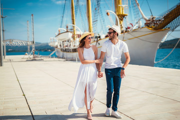 Happy young couple walking by the harbor of a touristic sea resort with sailboats on background