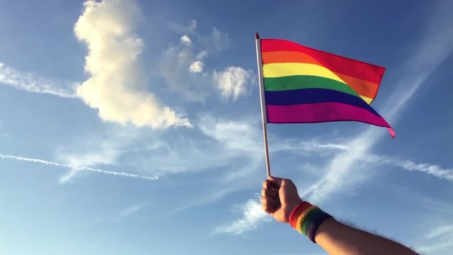 Hand wearing gay pride wrist band holding rainbow flag up in front of bright blue summer sky at sunset