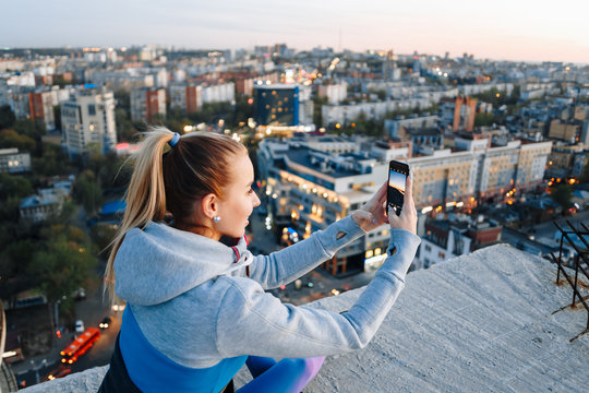 Young girl in sports uniform takes photos on the phone on the roof of a building over the city