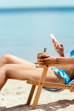 cropped image of woman holding cocktail in coconut shell and using smartphone while laying on deck chair on sandy beach