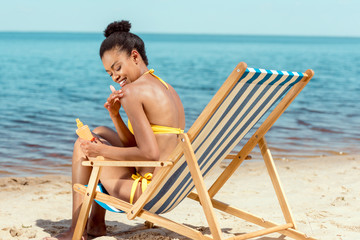smiling african american woman applying sunscreen lotion on skin while sitting on deck chair on sandy beach