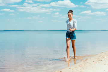 young smiling african american woman walking in sea water near sandy beach