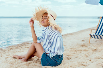 smiling african american woman in straw hat looking at camera while sitting on sandy beach with deck chair and beach umbrella