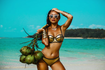 beautiful tanned girl in trendy bathing suit sunglasses stands with a bunch of coconuts in her hands on beach tropical island. Summer vacation, travel around the world, advertising swimwear new season