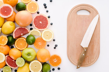 top view of citrus fruits with blueberries and wooden cutting board with knife on white surface