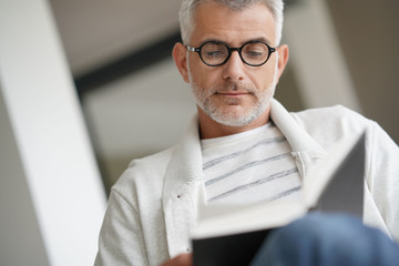 Trendy mature guy with eyeglasses reading book at home