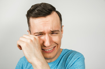 A young guy is crying wiping away tears, dressed in a blue T-shirt on a light background.