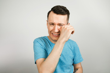 A young guy is crying wiping away tears, dressed in a blue T-shirt on a light background.