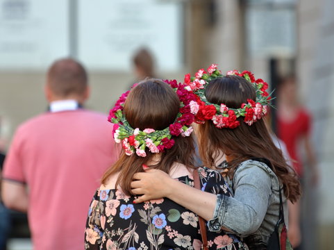 Dwie młode kobiety, długowłose, w kwietnych, roślinnych wiankach na głowach, stoją blisko, jedna obejmuje drugą ramieniem, w hippisowskich strojach, tło rozmyte