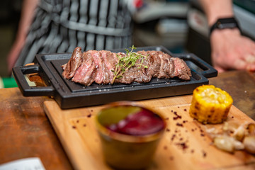 Cast iron plate with sliced grilled rib-eye steak on table with yellow potato wedges and oil in saucer over table