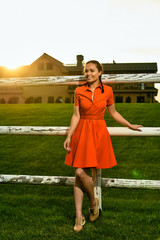 Beautiful brunette girl in a red dress in the village. Around green grass, a fence of logs and the sun sets. Grow birch. In the background is a large country house.