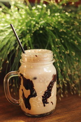 cold coffee frappe in glass jar with chocolate and foam in wooden table with fresh grass on...